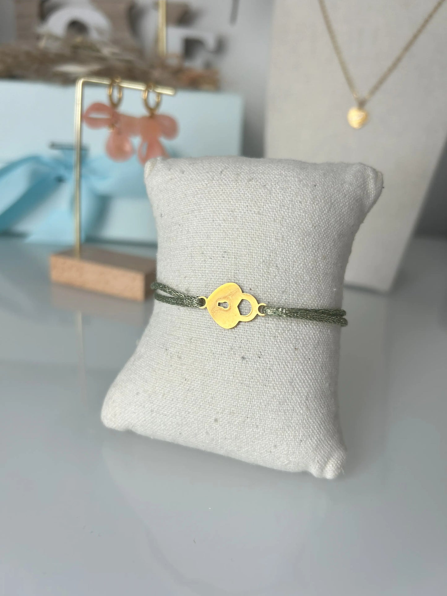 bracelet cordon scintillant vert à fil doré présenté sur un mini-coussin beige en coton.  Bijou orné d'un cadenas coeur en acier inoxydable doré. En fond un buste avec un collier doré et une paire de boucles d'oreilles dorées et saumon sur un présentoir en bambou. 