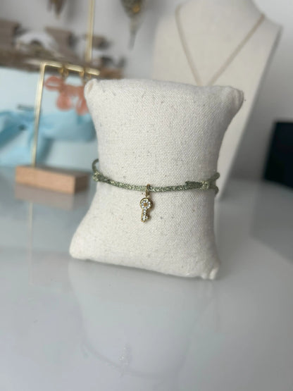 bracelet cordon scintillant vert olive à fil doré présenté sur un mini-coussin beige en coton.  Bijou orné d'une clé avec strass en acier inoxydable doré. En fond un buste avec un collier doré et une paire de boucles d'oreilles dorées et saumon sur un présentoir en bambou. 
