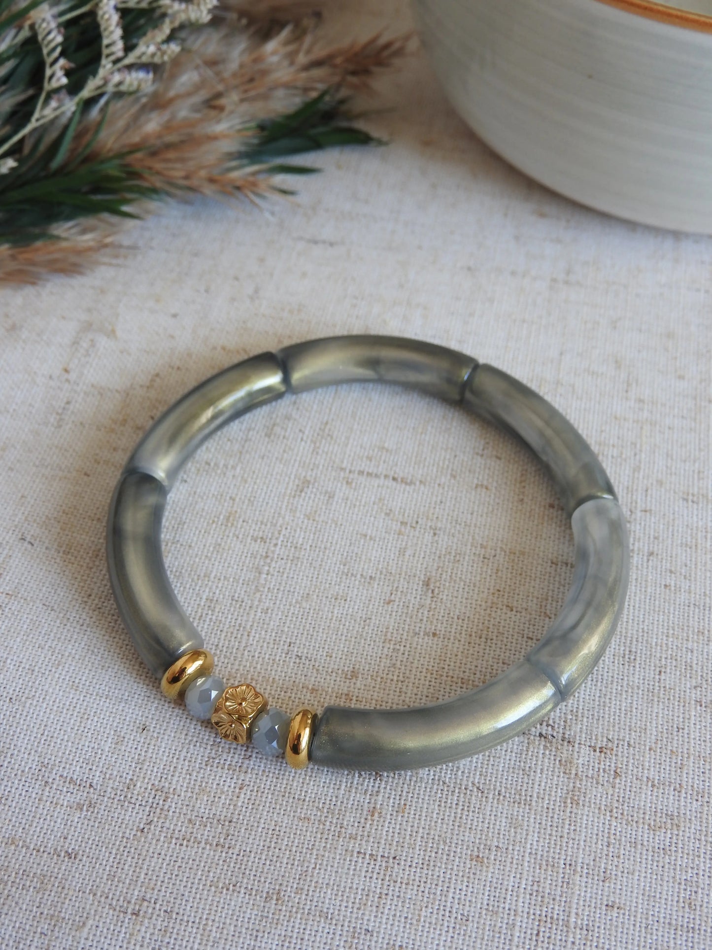 Bracelet jonc à perles tube gris irisées et orné de perles de verre grises et des perles rondelles et une perle cube au motif fleuri en acier inoxydable doré. Bijou posé sur une nappe beige décorée d'une brindille et un bol en céramique.