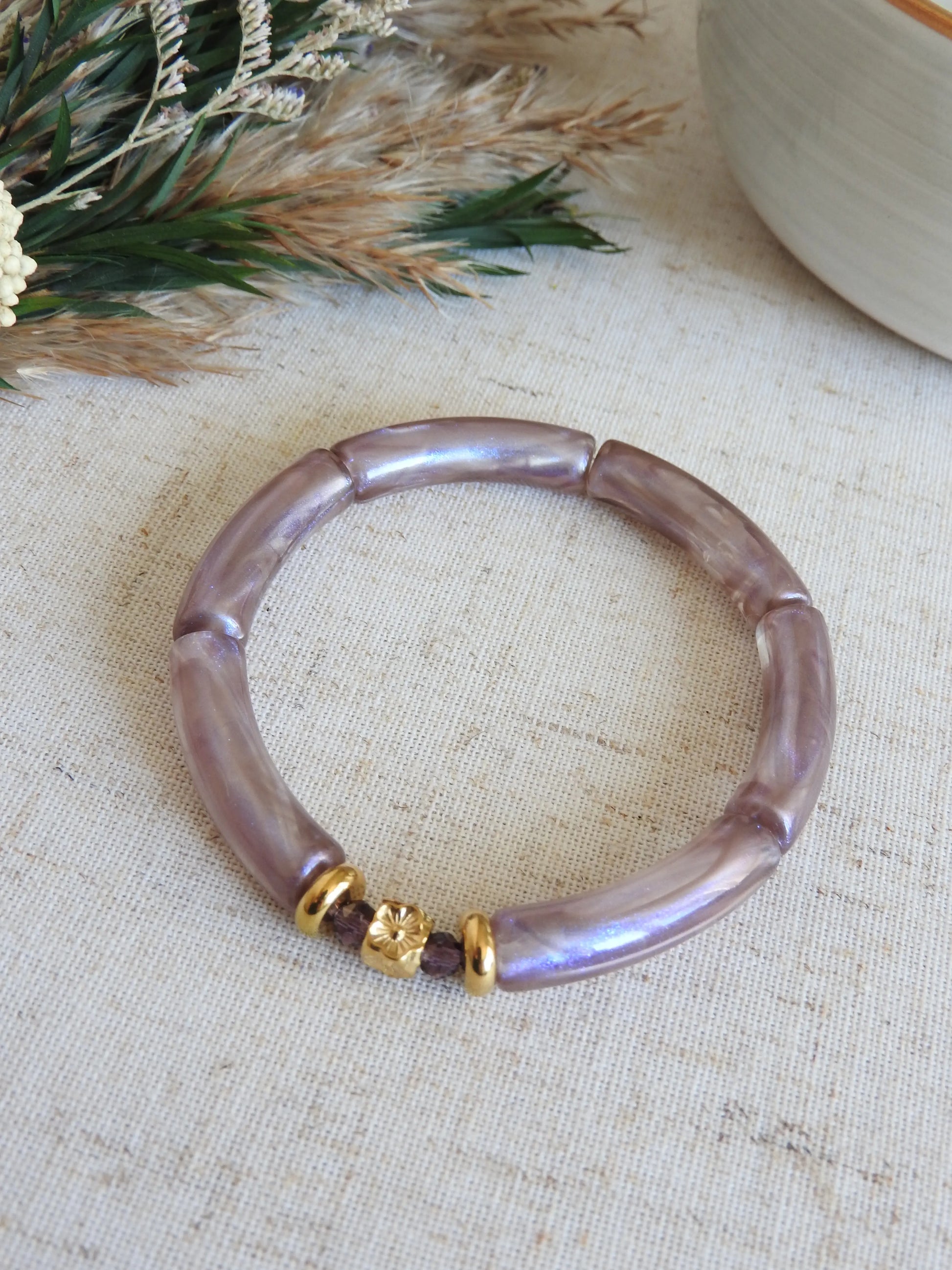 Bracelet jonc à perles tube parme irisées et orné de perles de verre violet et des perles rondelles et une perle cube au motif fleuri en acier inoxydable doré. Bijou posé sur une nappe beige décorée d'une brindille et un bol en céramique.