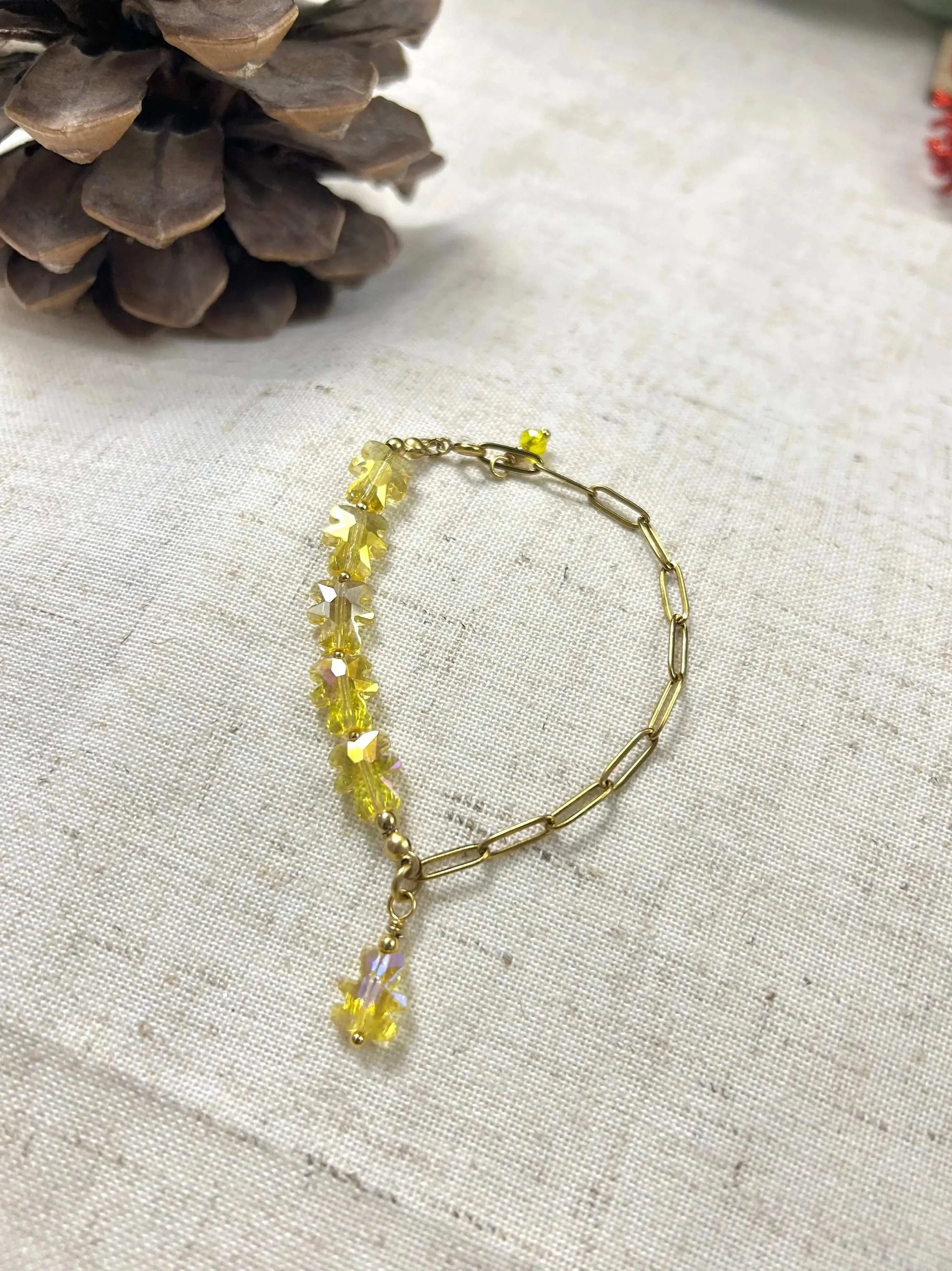 bracelet mi-chaîne rectangle en acier inoxydable doré et mi-perles de verre en forme d'ours jaune. Bijou posé à plat sur une nappe beige en coton décorée de brindilles.