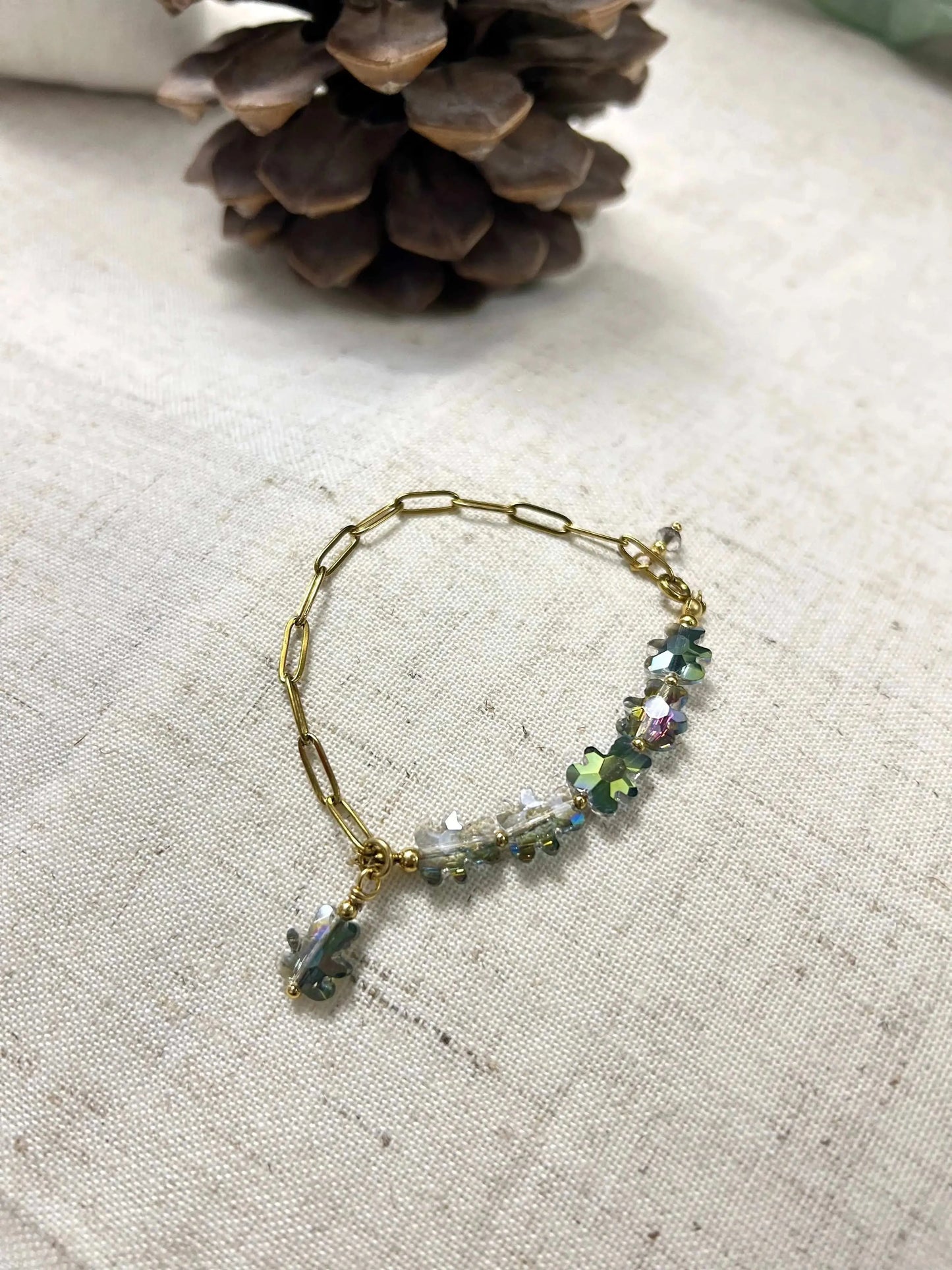 bracelet mi-chaîne rectangle en acier inoxydable doré et mi-perles de verre en forme d'ours multicouleurs. Bijou posé à plat sur une nappe beige en coton décorée d'une pomme de pin.