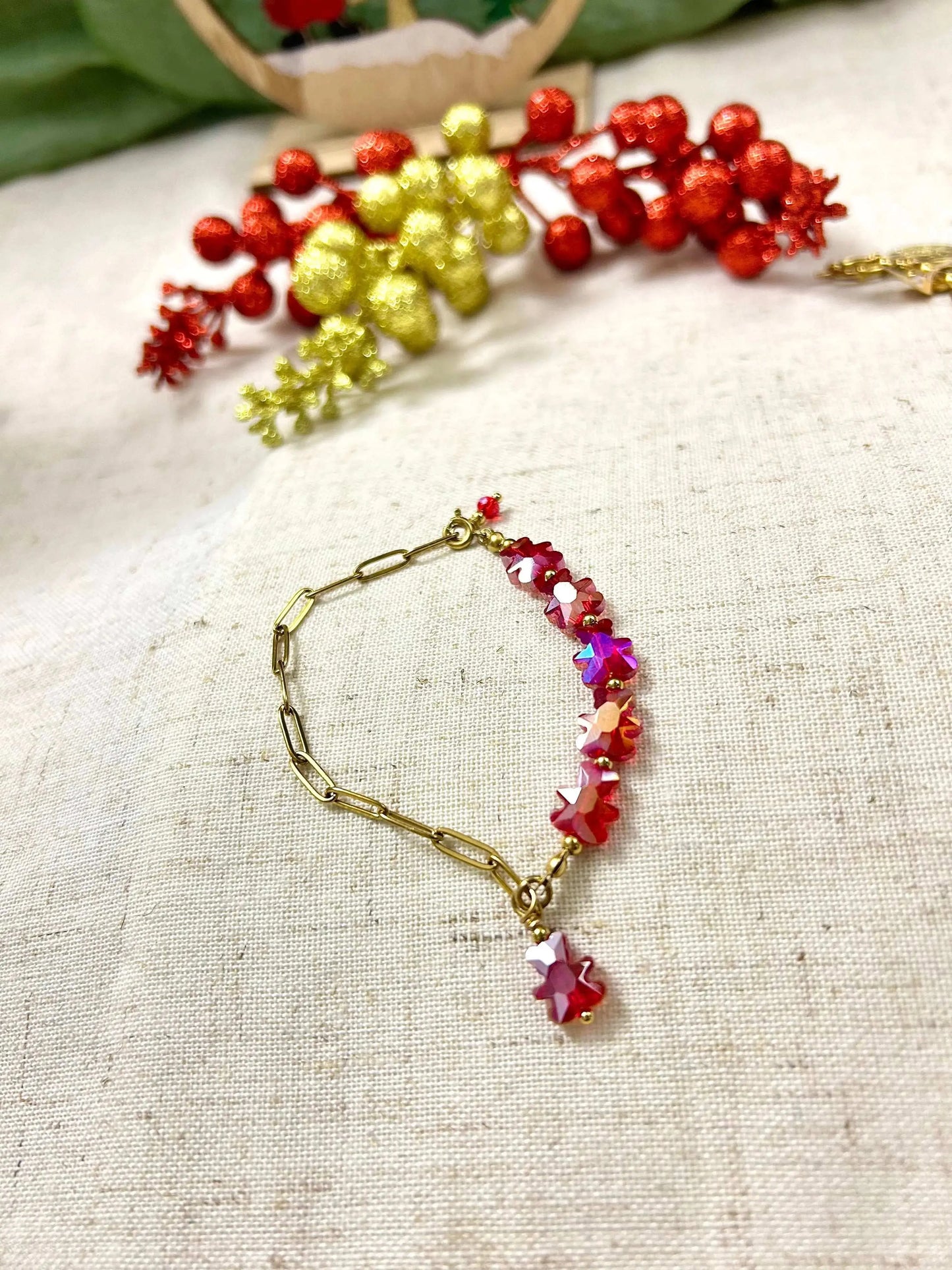 bracelet mi-chaîne rectangle en acier inoxydable doré et mi-perles de verre en forme d'ours rouge. Bijou posé à plat sur une nappe beige en coton décorée de décoration de noël.