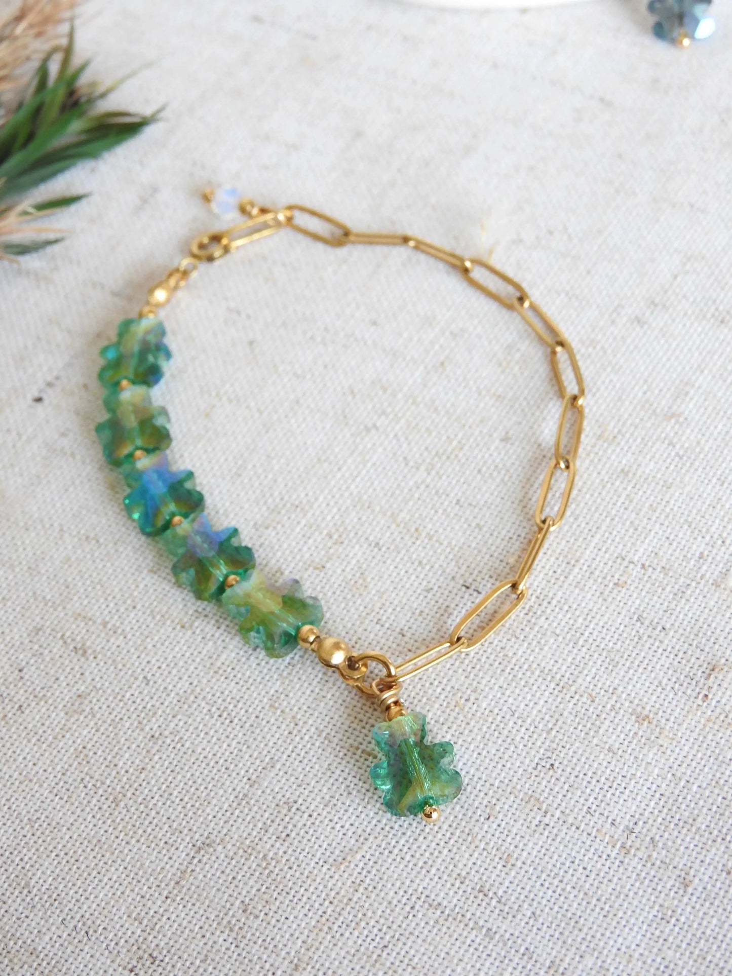 bracelet mi-chaîne rectangle en acier inoxydable doré et mi-perles de verre en forme d'ours vert. Bijou posé à plat sur une nappe beige en coton décorée de brindilles.
