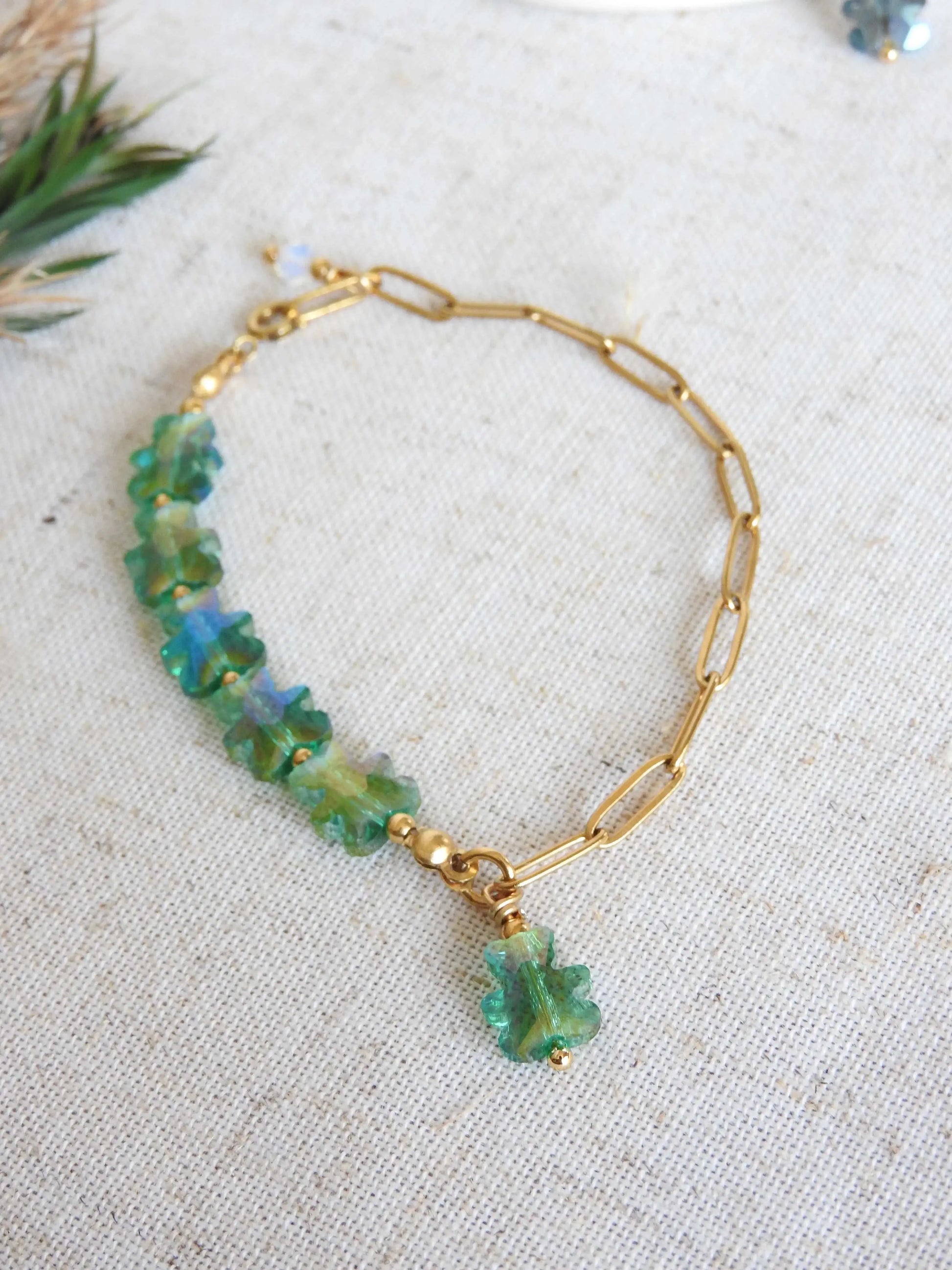 bracelet mi-chaîne rectangle en acier inoxydable doré et mi-perles de verre en forme d'ours vert. Bijou posé à plat sur une nappe beige en coton décorée de brindilles.