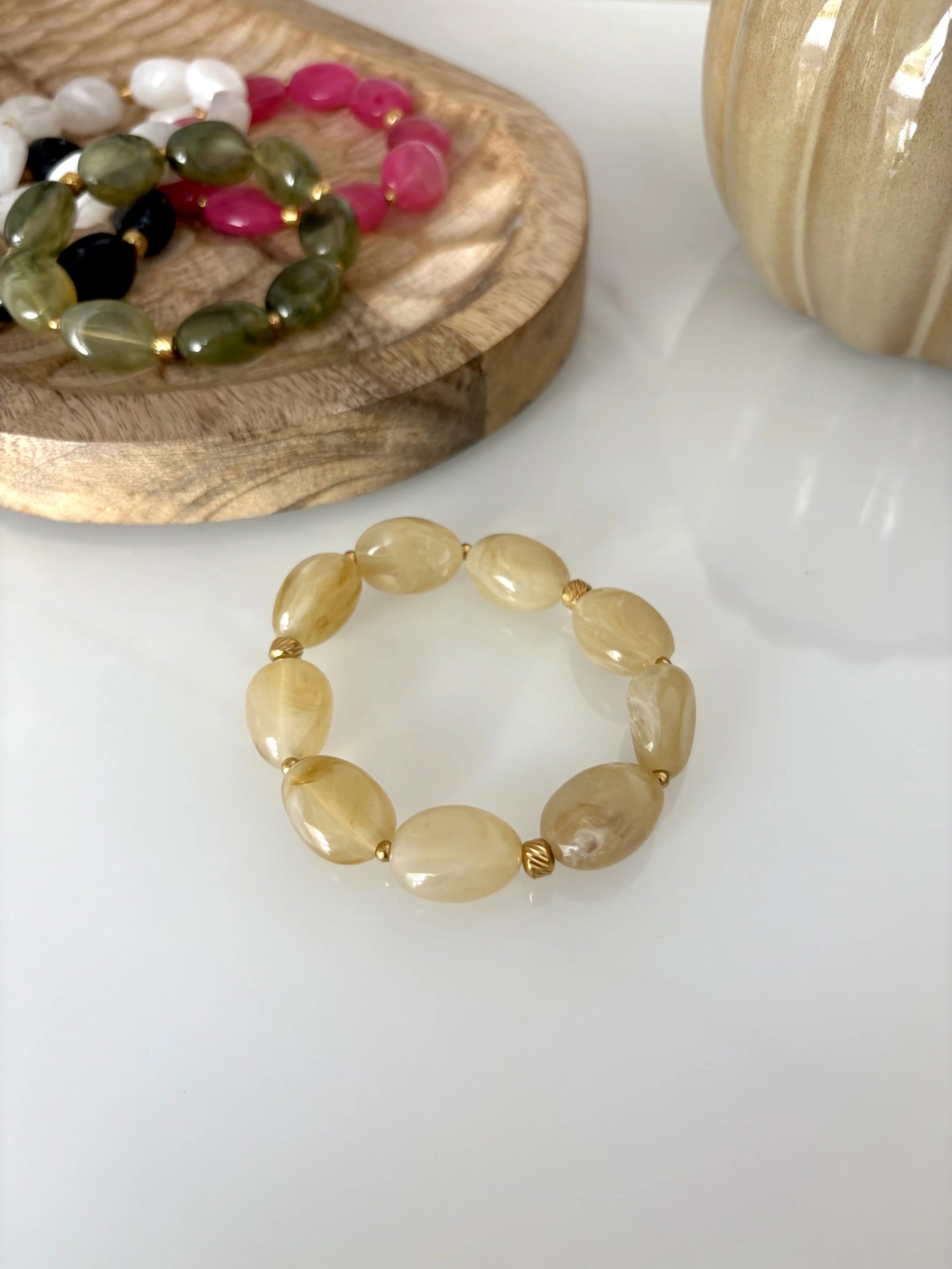 Bracelet élastique à grandes perles colorées kaki et orné de petites perles dorées en acier inoxydable. Bijou posé sur une table en verre trempée et décorée d'un plateau en bambou et des bracelets du même modèle de coloris rose, blanc, noir, rose et vert olive.