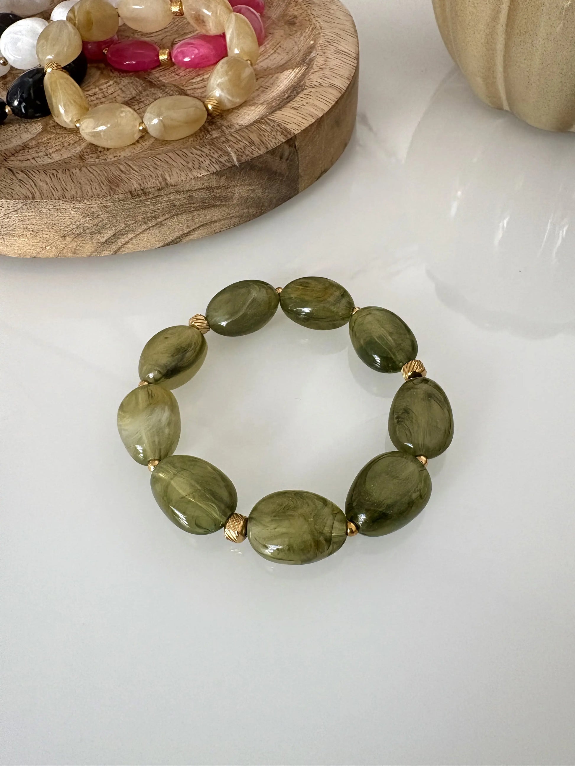 Bracelet élastique à grandes perles colorées vert olive et orné de petites perles dorées en acier inoxydable. Bijou posé sur une table en verre trempée et décorée d'un plateau en bambou et des bracelets du même modèle de coloris rose, blanc, noir et rose.