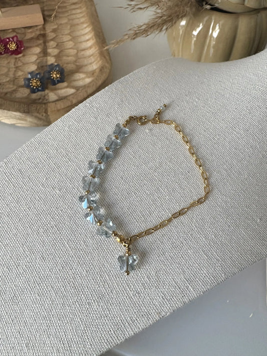 Bracelet mi chaine rectangle en acier inoxydable doré et perles de verre en forme de papillon bleu-gris. Bijou présenté sur un support beige avec en fond un plateau en bambou avec des boucles d'oreilles fleur hibiscus framboise et bleu foncé 