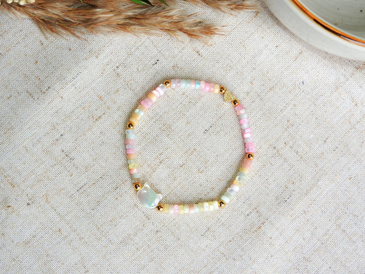 bracelet perles rondelles heishi en nacre teintées de tons pastels, orné d'une perle chat en nacre blanche. Bijou posé sur une nappe beige.
