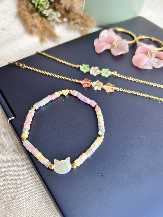 Bracelet en perles rondelles heishi en nacre teintées, orné d'une perle chat en nacre blanche. Bijou posé sur un pc portable avec d'autres bijoux : bracelets chaine et perles en étoile et boucles d'oreilles créoles avec pétales couleur rose marbré.