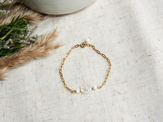 bracelet chaîne maille rectangle orné d'une perle chat en nacre blanche. Bijou posé sur une nappe beige décorée d'un mini-bouquet de fleurs.