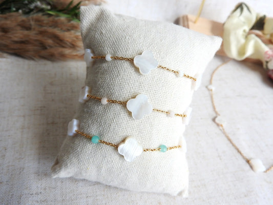 trois bracelets chaînes en acier inoxydable doré perlés de perles de verre turquoise, rose poudre et blanc ainsi que des perles trèfle en nacre blanche. Bijoux présentés sur un mini-coussin beige en coton posé sur une table décorée de brindilles. 
