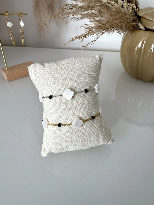 deux bracelets chaînes en acier inoxydable doré et argenté perlés de perles noires et trèfle en nacre blanche. Bijoux présentés sur un mini-coussin beige en coton posé sur une table décorée de brindilles, un pot en céramique kaki et des boucles d'oreilles pendantes.