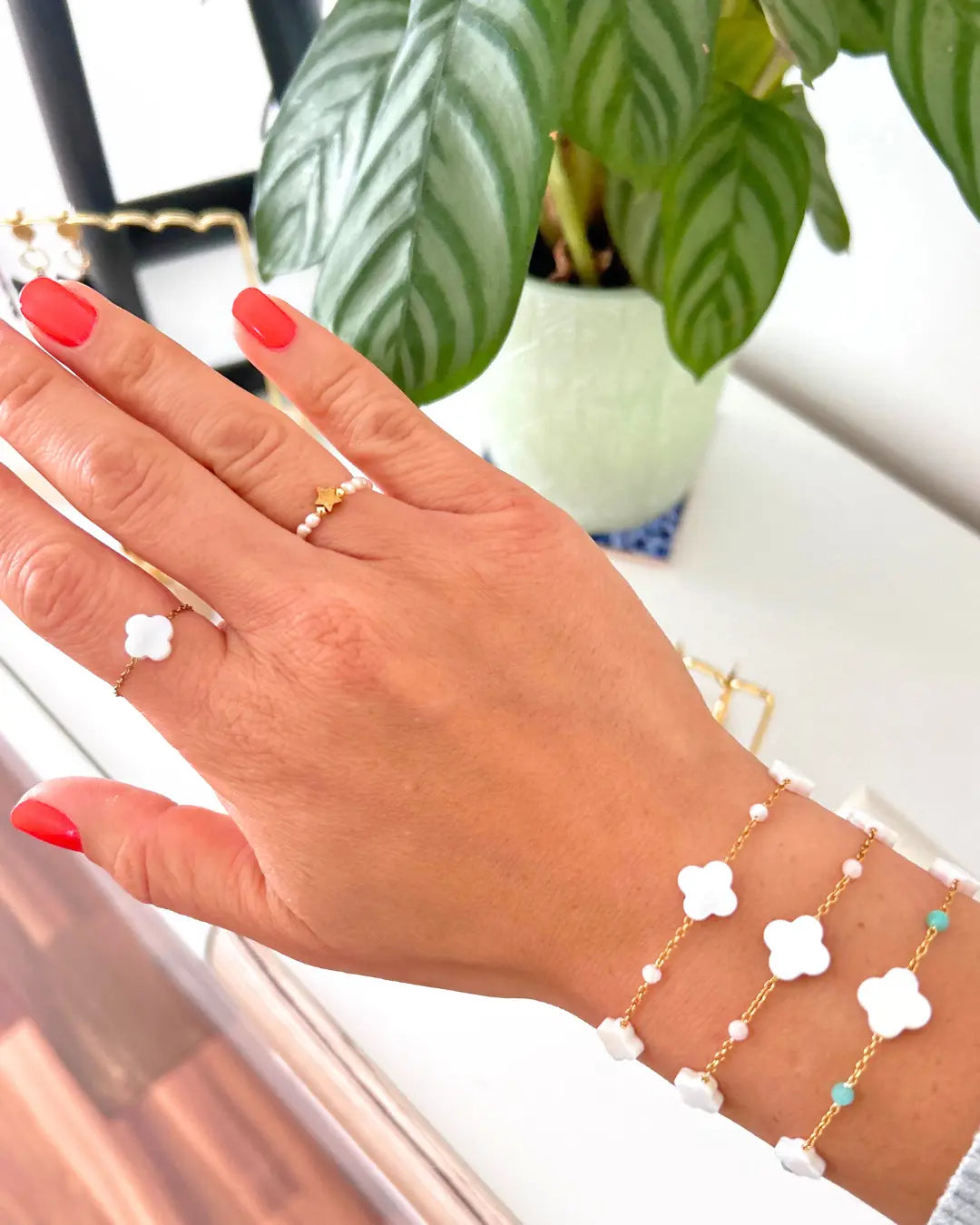 trois bracelets chaînes en acier inoxydable doré perlés en verre turquoise, rose poudre et blanc ainsi que des perles trèfle en nacre blanche. Bijoux portés au poignet avec une bague en perles d'eau douce blanche et une perle étoile en acier doré à l'annulaire et une autre bague chaînette avec une perle trèfle en nacre blanche à l'index.