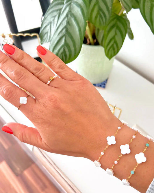 trois bracelets chaînes en acier inoxydable doré perlés en verre turquoise, rose poudre et blanc ainsi que des perles trèfle en nacre blanche. Bijoux portés au poignet avec une bague en perles d'eau douce blanche et une perle étoile en acier doré à l'annulaire et une autre bague chaînette avec une perle trèfle en nacre blanche à l'index.