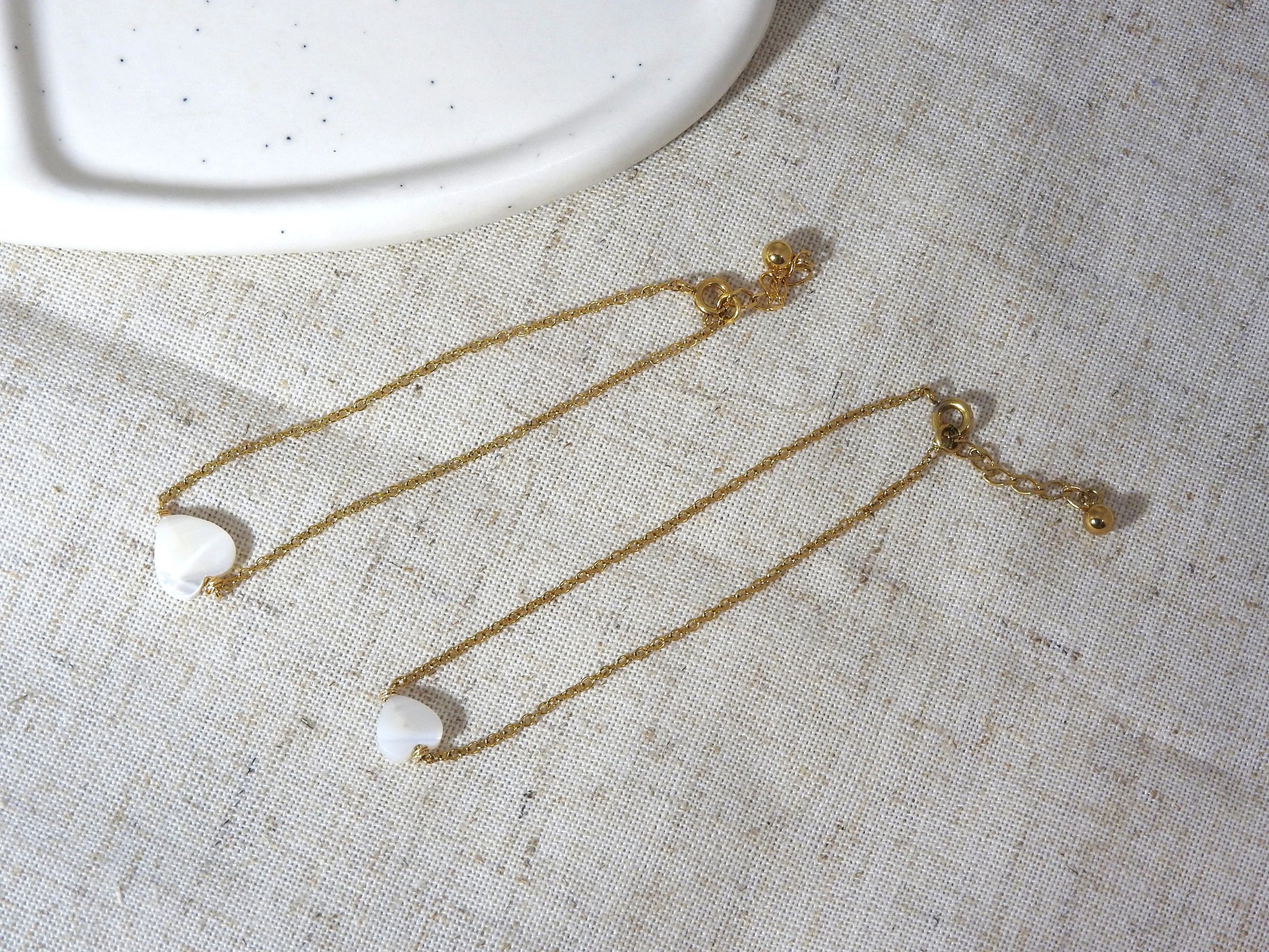 Bracelets chaîne fine en acier inoxydable doré posés à plat sur une nappe beige en coton. Bijoux ornés d'une perle coeur en nacre blanche.