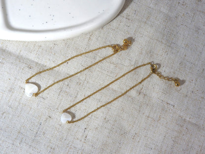 Bracelets chaîne fine en acier inoxydable doré posés à plat sur une nappe beige en coton. Bijoux ornés d'une perle coeur en nacre blanche.