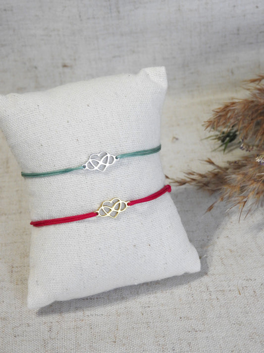 deux bracelets cordon en nylon coloré turquoise et fuchsia, ornés d'un coeur infini en acier inoxydable doré et argenté. Bijoux présentés sur un mini-coussin beige en coton posés sur une table décorée d'une brindille.