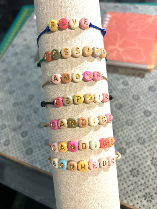 Bracelets cordon réglable multicouleurs avec un mot personnalisé tel que rêve, passion, amour, espoir, bonheur. Bijoux avec des perles rondes plates en acrylique colorées. Bijoux posés sur un présenoir à bracelet beige. 