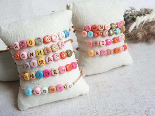 Bracelets cordon réglable couleur champagne avec un mot personnalisé tel que adorable, bonheur, aimante, lumineuse. Bijou avec des perles rondes plates en acrylique colorées. Bijoux posés sur un mini-coussin beige, posé une nappe décorée de brindilles et bol en céramique.