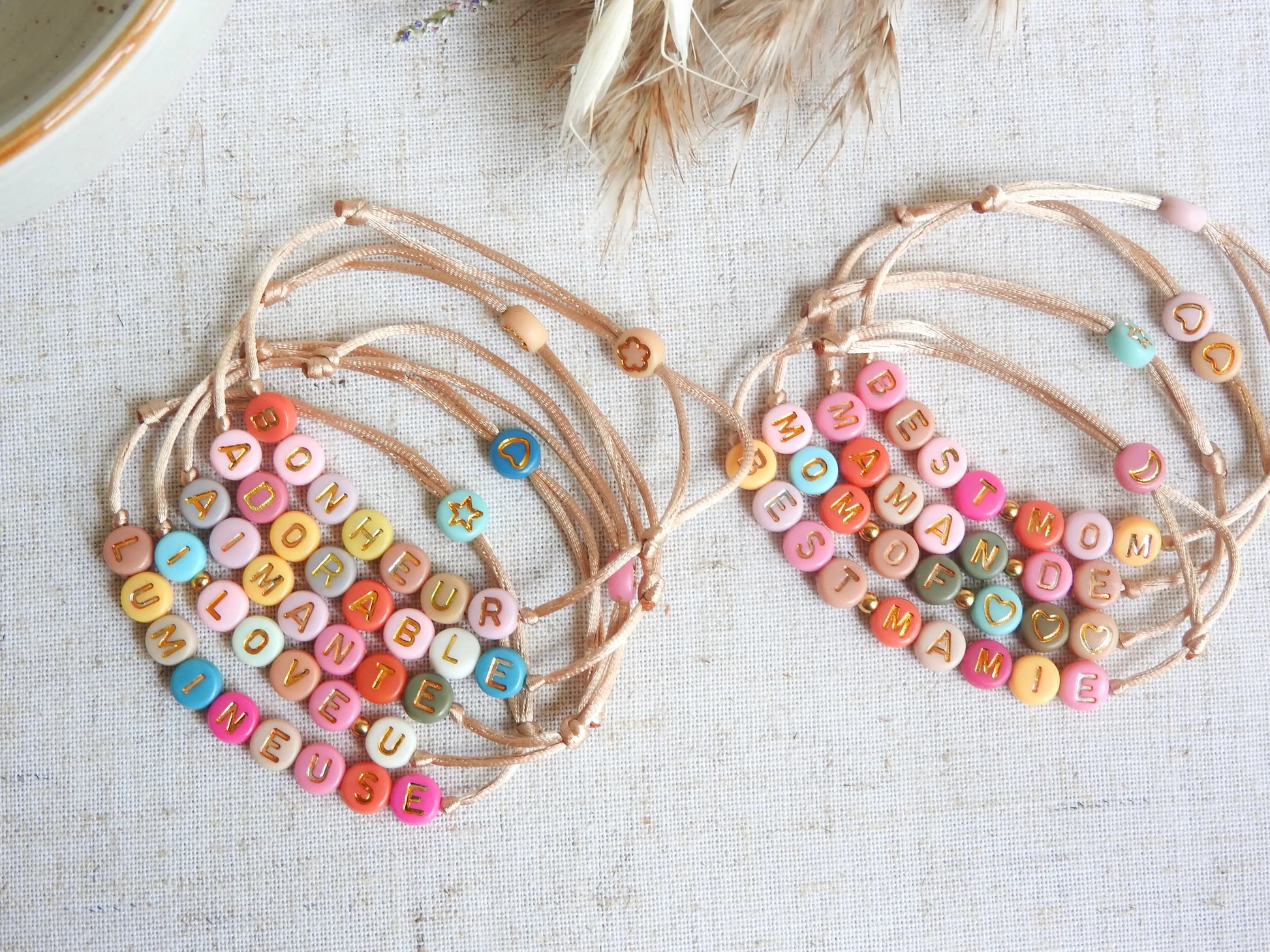Bracelets cordon réglable couleur champagne avec un mot personnalisé tel que adorable, bonheur, aimante, lumineuse. Bijoux avec des perles rondes plates en acrylique colorées. Bijoux posés sà plat sur une nappe beige décorée de brindilles.