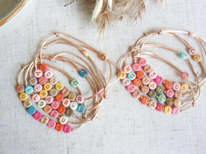 Bracelets cordon réglable couleur champagne avec un mot personnalisé tel que adorable, bonheur, aimante, lumineuse. Bijoux avec des perles rondes plates en acrylique colorées. Bijoux posés sà plat sur une nappe beige décorée de brindilles.