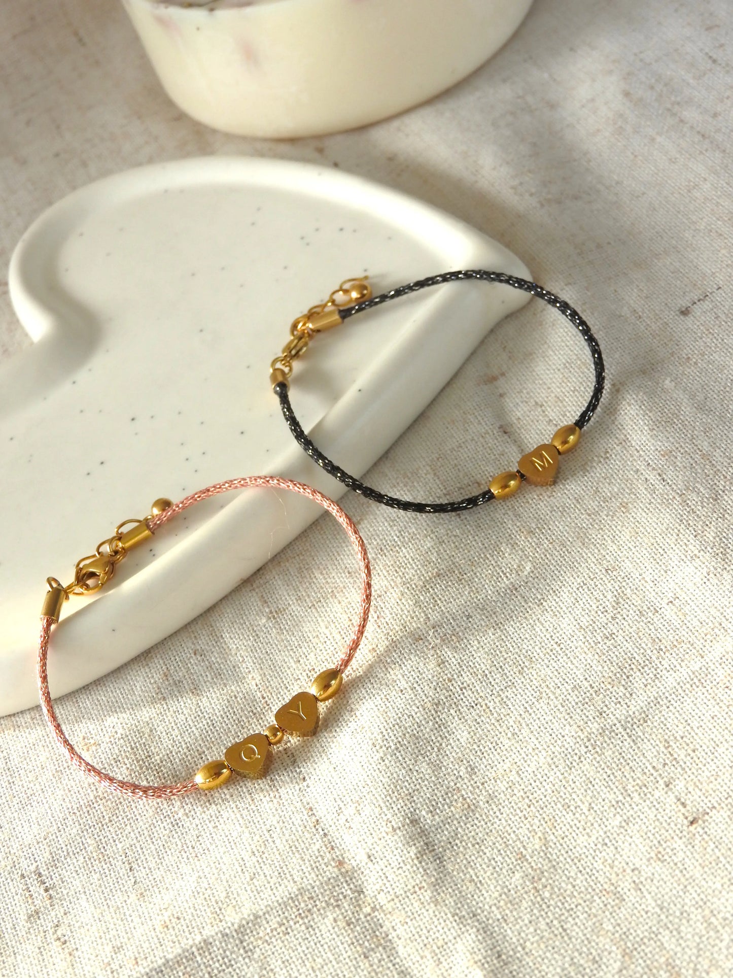 Bracelets cordon noir et saumon scintillant ornés de 3 perles dont une en forme de coeur plat gravée de la lettre M pour le bracelet noir et les lettres Q et Y pour le bracelet saumon en acier inoxydable doré. Bijoux posés sur une coupelle blanche en céramique.