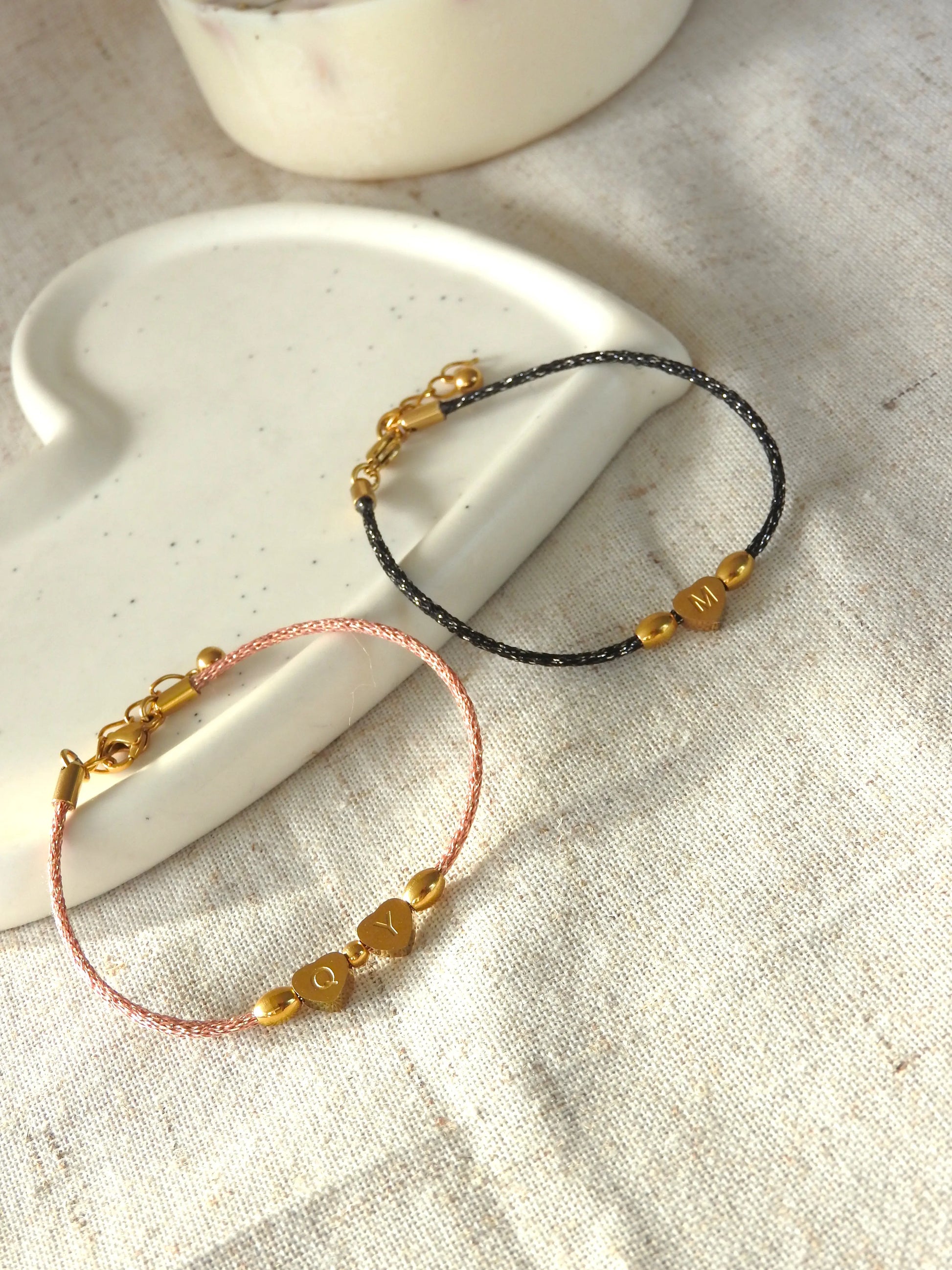 Bracelets cordon noir et saumon scintillant ornés de 3 perles dont une en forme de coeur plat gravée de la lettre M pour le bracelet noir et les lettres Q et Y pour le bracelet saumon en acier inoxydable doré. Bijoux posés sur une coupelle blanche en céramique.