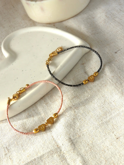 Bracelets cordon noir et saumon scintillant ornés de 3 perles dont une en forme de coeur plat gravée de la lettre M pour le bracelet noir et les lettres Q et Y pour le bracelet saumon en acier inoxydable doré. Bijoux posés sur une coupelle blanche en céramique.