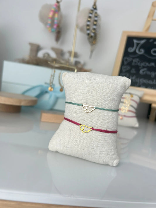 Deux bracelets cordon en nylon tressé turquoise et fuschia présentés sur un mini-coussin en coton beige. Bijoux ornés d'un intercalaire en aile d'ange en acier inoxydable doré. Bijoux posés sur une table en verre trempée avec en fond des porte-clés. 