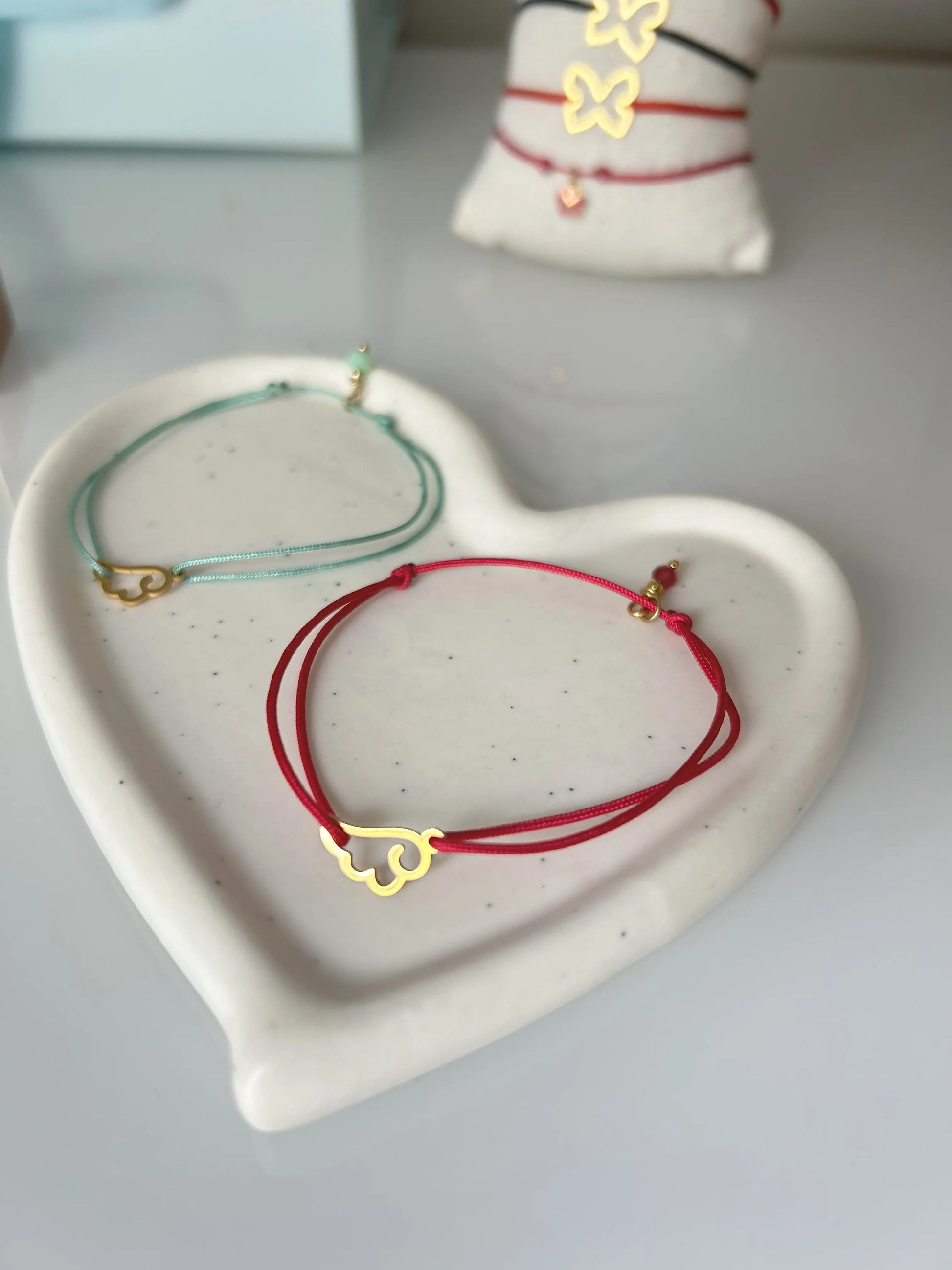 Deux bracelets cordon en nylon tressé turquoise et fuchsia présentés sur une coupelle coeur en céramique blanche. Bijoux ornés d'un intercalaire en aile d'ange et pampille en acier inoxydable doré.