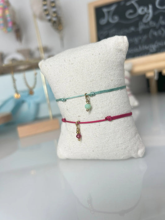 Deux bracelets cordon en nylon tressé turquoise et fuchsia présentés sur un mini-coussin en coton beige. Bijoux ornés d'une pampille colorée en perle de verre et en acier inoxydable doré. Bijoux posés sur une table en verre trempée avec en fond des porte-clés. 