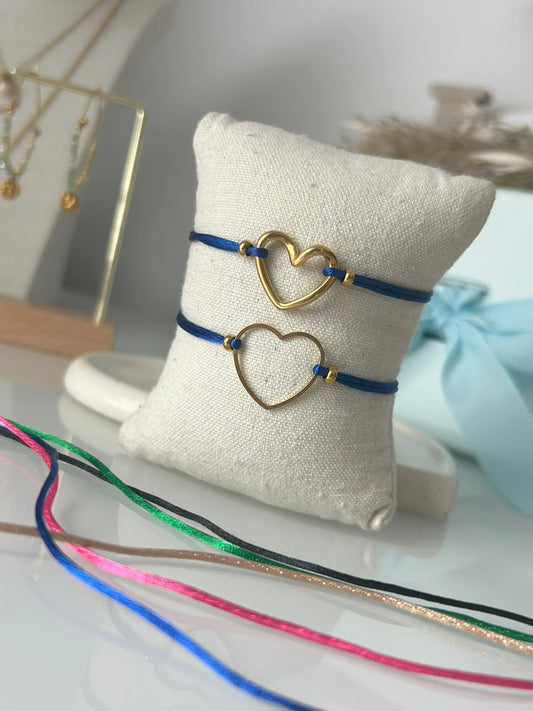 bracelets cordon ajustable en nylon bleu roi ornés d'un cœur lisse et un coeur courbé en acier inoxydable doré. Bijoux présentés sur un mini-coussin beige en coton, posé sur une table décorée de cordon en nylon aux couleurs noir, vert électrique, champagne, rose fuchsia et bleu roi.