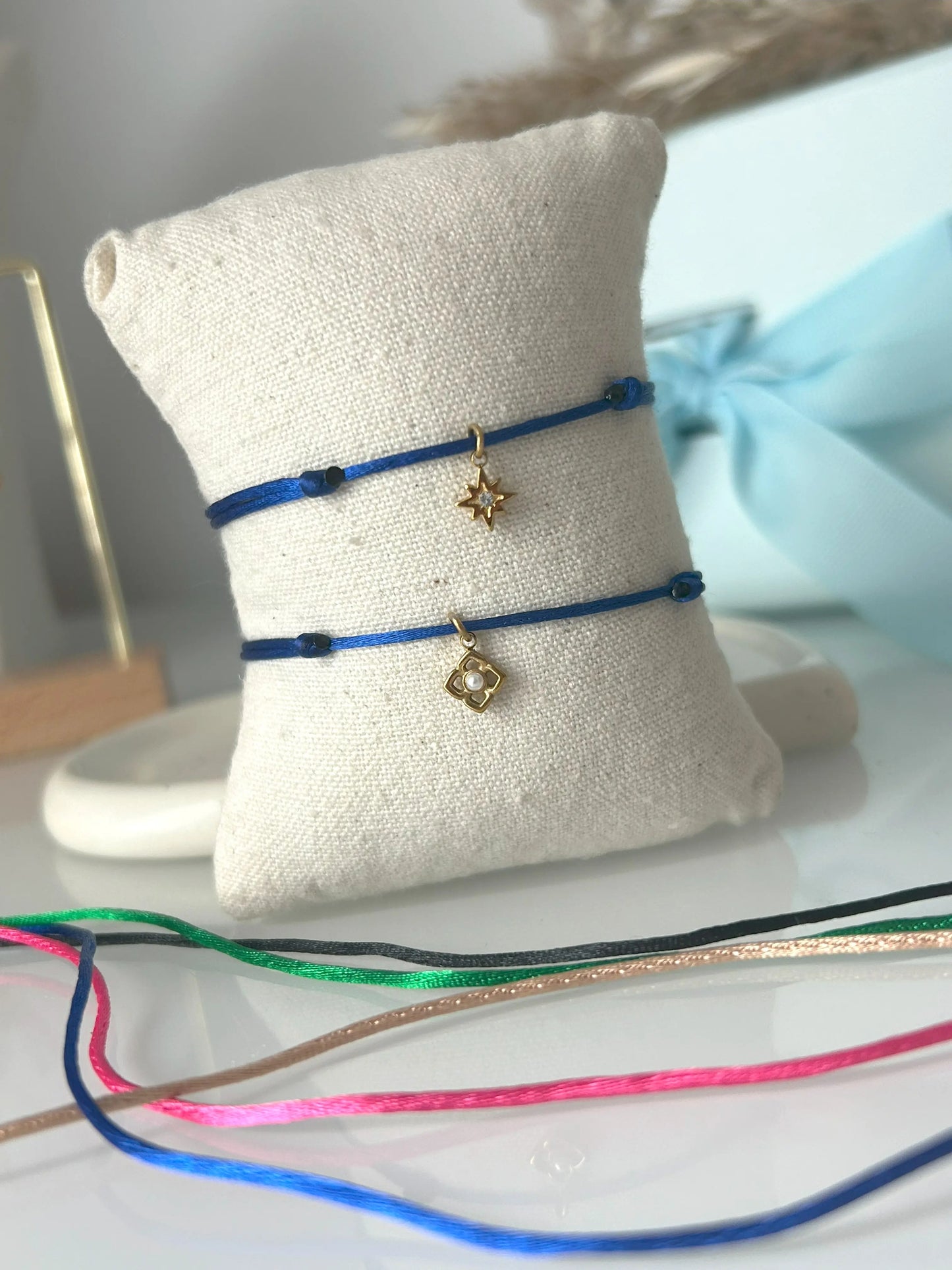 bracelets cordon ajustable en nylon bleu roi ornés d'une pampille étoile du nord avec une pierre en oxyde de zirconium et une pampille fleur avec perle blanche imitation perle d'eau douce et en acier inoxydable doré. Bijoux présentés sur un mini-coussin beige en coton, posé sur une table décorée de cordon en nylon aux couleurs noir, vert électrique, champagne, rose fuchsia et bleu roi.