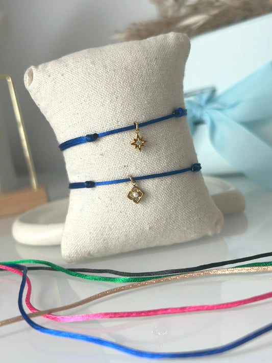 bracelets cordon ajustable en nylon bleu roi ornés d'une pampille étoile du nord avec une pierre en oxyde de zirconium et une pampille fleur avec perle blanche imitation perle d'eau douce et en acier inoxydable doré. Bijoux présentés sur un mini-coussin beige en coton, posé sur une table décorée de cordon en nylon aux couleurs noir, vert électrique, champagne, rose fuchsia et bleu roi.