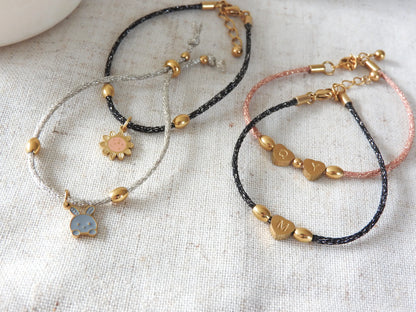 Bracelets à cordon noir, saumon et gris scintillants ornés de perles en forme d'olive et en forme de coeur gravées de lettres M, Q et Y. D'autres bracelets à breloques tournesol et lapin. Bijoux posés sur une nappe beige en coton et acier inoxydable doré.