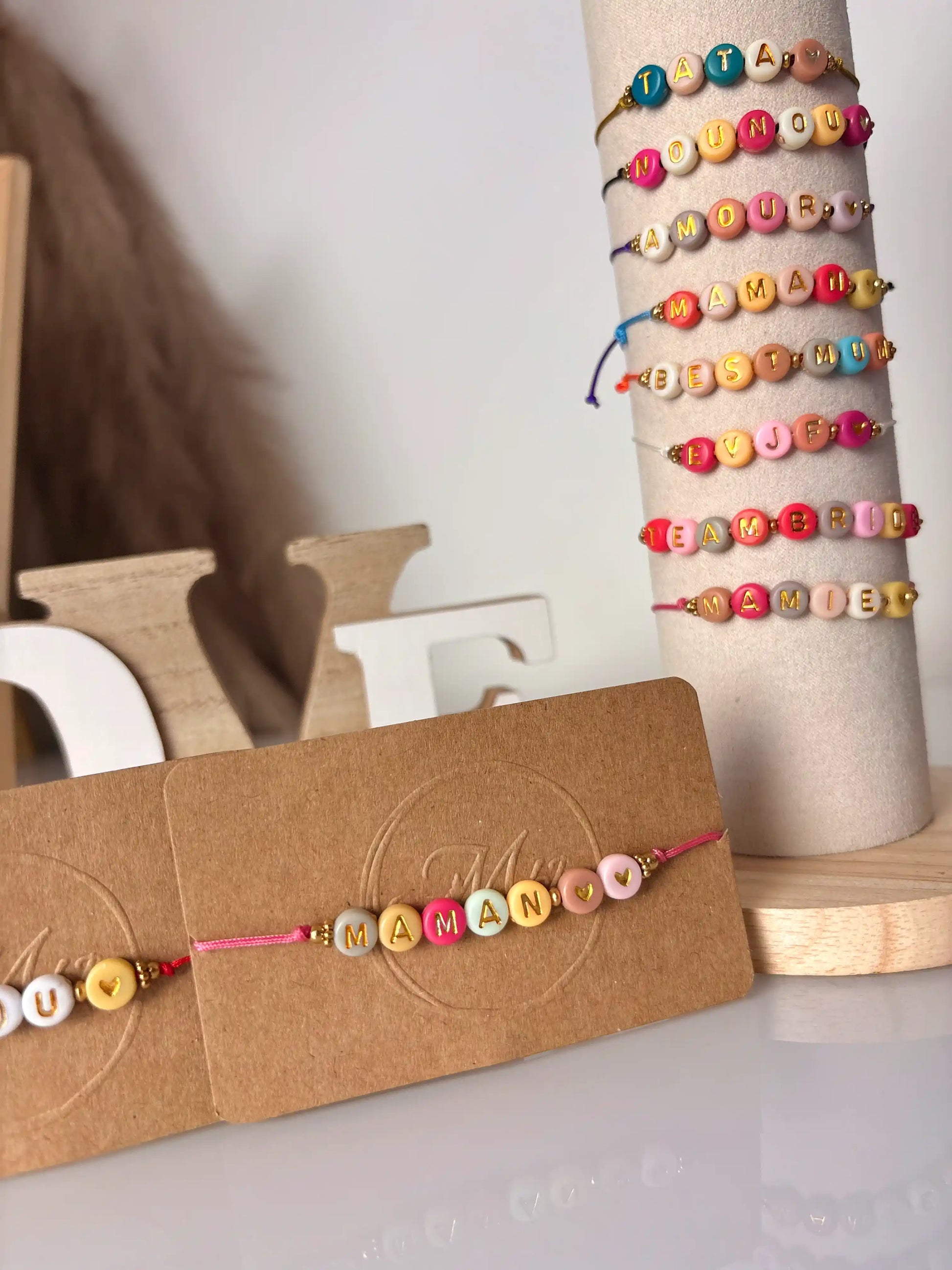 Bracelets à cordon coloré avec un message en perles colorées à personnaliser. Bijoux présentés sur un support tube vertical et un carton kraft.