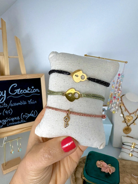 bracelets cordon scintillant à fil argenté de coloris noir, vert olive et saumon, ornés d'un cadenas coeur en acier inoxydable doré et une pampille à l'arrière clé en oxydes de zirconium. Bijoux présentés sur un mini-coussin de présentation tenu par une main. 