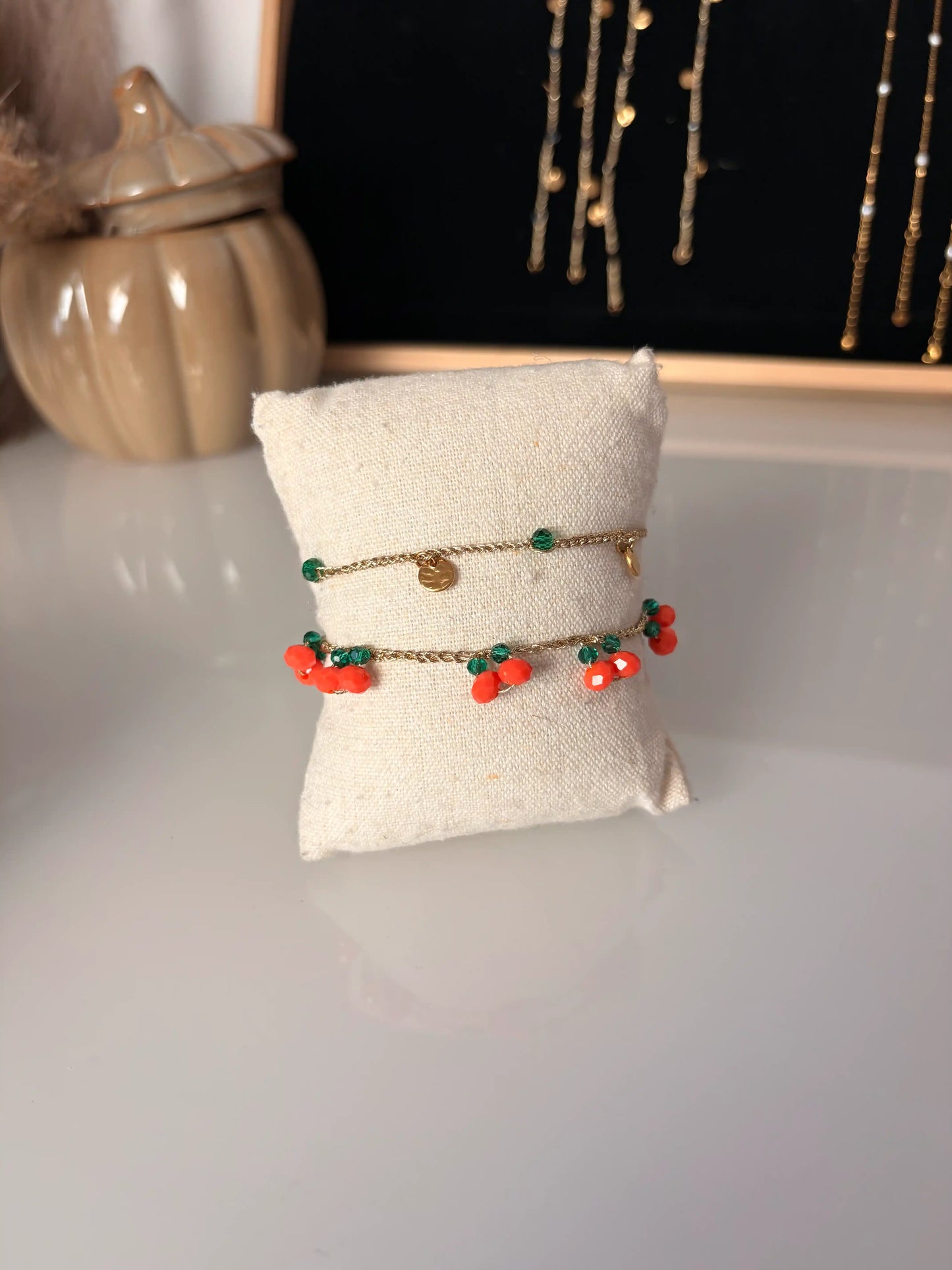 Bracelets à fil crochetés doré présentés sur un mini coussin en coton beige. Bracelets ornés de perles de verre orange et vert émeraude formant des oranges sur le bracelet et des petits sequins ronds martelés en acier inoxydable doré.