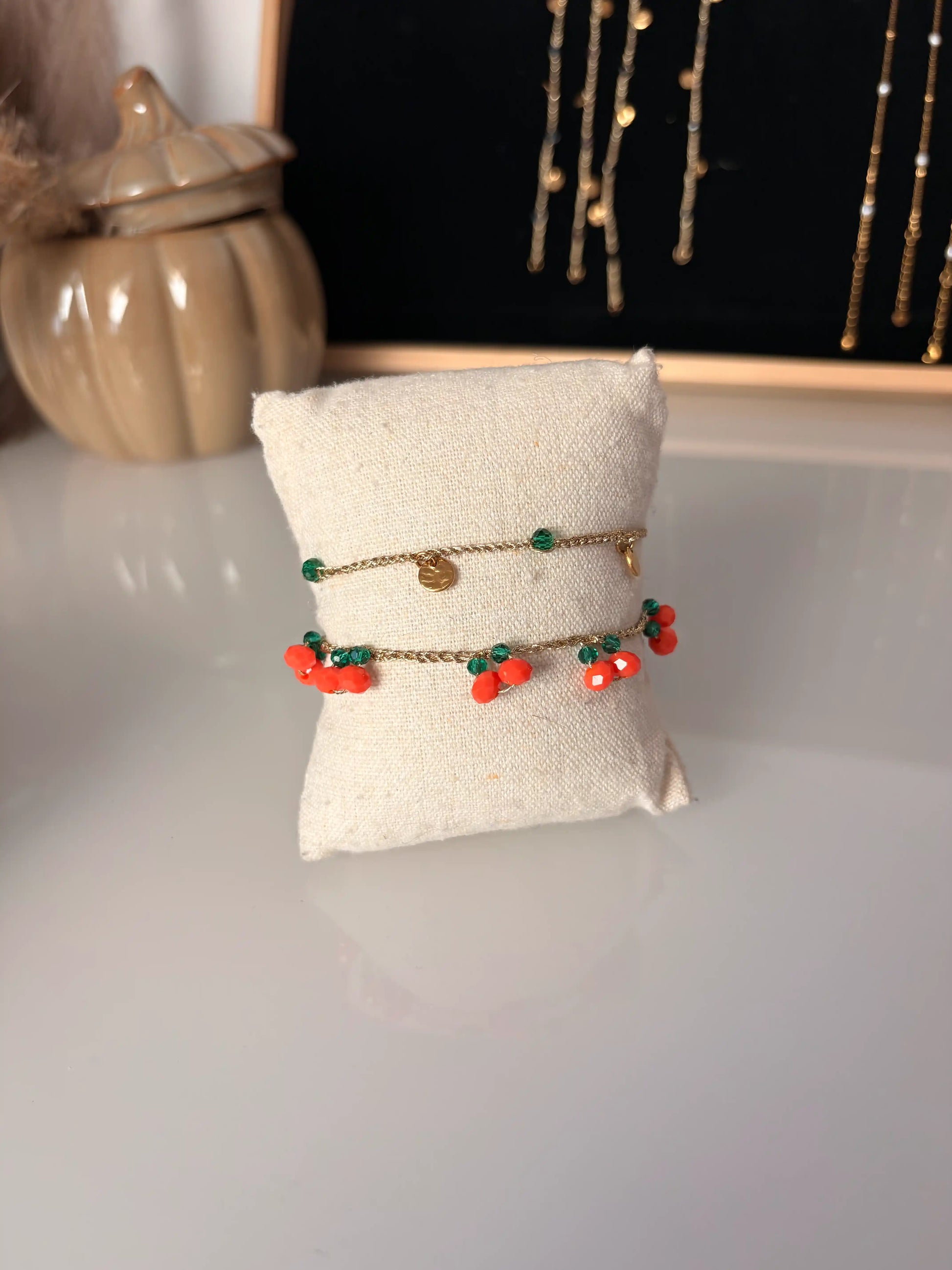 Bracelets à fil crochetés doré présentés sur un mini coussin en coton beige. Bracelets ornés de perles de verre orange et vert émeraude formant des oranges sur le bracelet et des petits sequins ronds martelés en acier inoxydable doré.