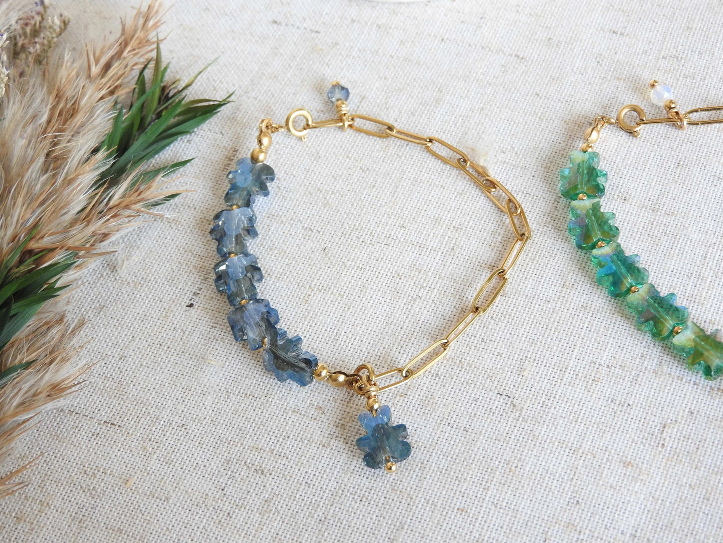 deux bracelets posés à plat sur une nappe beige en coton décorée d'un mini-bouquet de fleurs. Bracelets mi-chaîne à maille rectangle en acier inoxydable doré et mi-perles de verre en forme d'ours colorées bleu nuit et vert.