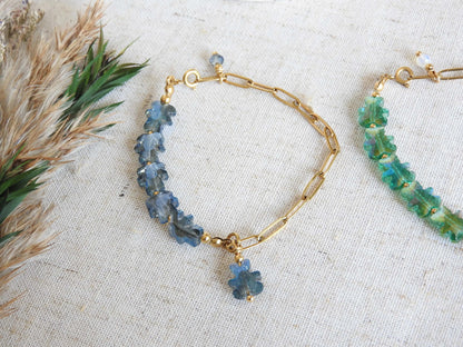 deux bracelets posés à plat sur une nappe beige en coton décorée d'un mini-bouquet de fleurs. Bracelets mi-chaîne à maille rectangle en acier inoxydable doré et mi-perles de verre en forme d'ours colorées bleu nuit et vert.