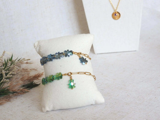 deux bracelets présentés sur un mini-coussin beige en coton disposé sur une nappe beige décorée d'un mini-bouquet de fleurs. Bracelets mi-chaîne à mailles rectangle en acier inoxydable doré et mi-perles en forme d'ours en verre et colorées bleu gris et vert. 