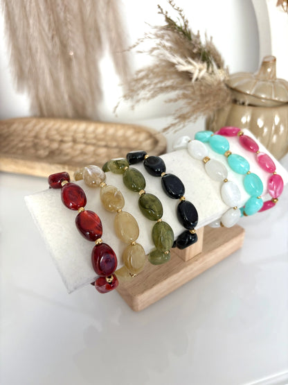 Bracelets à grandes perles colorées et alignés sur un présentoir en bambou. sept bracelets de couleurs bordeaux, kaki, vert olive, noir, blanc, turquoise et rose. Présentoir sur une table en verre trempée et décorée d'une citrouille en céramique et plateau en bois.