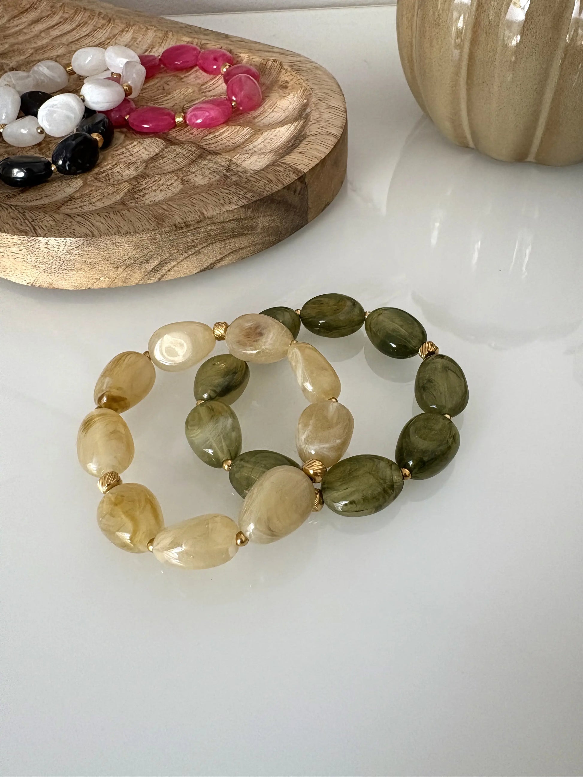 Bracelets élastique à grandes perles colorées kaki et vert olive et ornés de petites perles dorées en acier inoxydable. Bijoux posés sur une table en verre trempée et décorée d'un plateau en bambou et des bracelets du même modèle de coloris rose, blanc, noir.