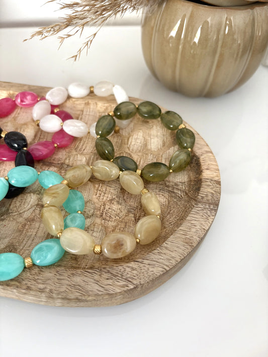 Bracelets superposés sur un plateau en bambou avec de grandes perles plates irrégulières en acrylique et de couleurs turquoise, blanc, rose, noir, kaki et olive ainsi que des perles en acier inoxydable doré.