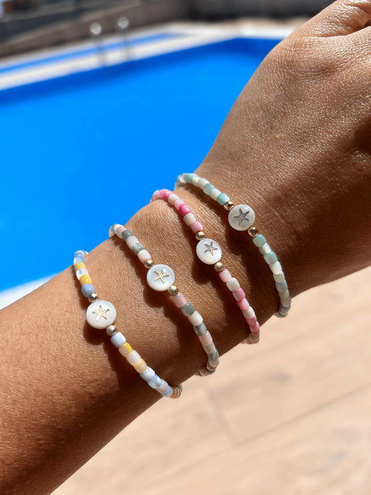 Bracelets perlés en nacre colorée bleu, bleu-jaune, rose-blanc et rose-gris, ornés d'une perle à motif étoile de mer en nacre blanche. Bijoux portés eau poignet sur fond d'une piscine extérieure.