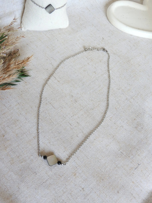 Collier chaine en acier inoxydable argenté posé à plat sur une nappe beige. Bijou orné d'une perle carré en nacre aux reflets noir, gris et blanc, et perles de verre.