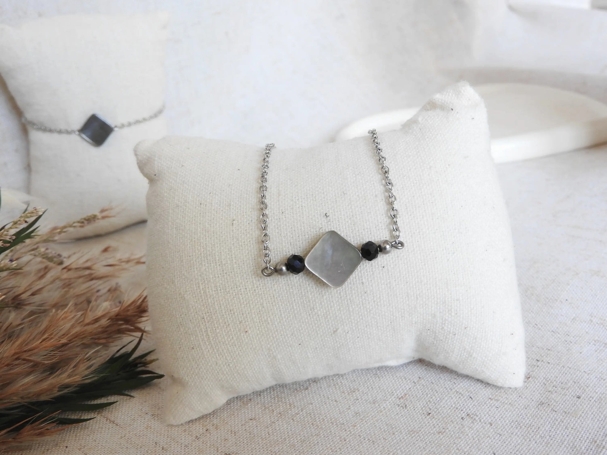 Collier chaîne en acier inoxydable argenté posé sur un mini-coussin beige. Bijou orné d'une perle en nacre aux reflets gris, noir et blanc. 