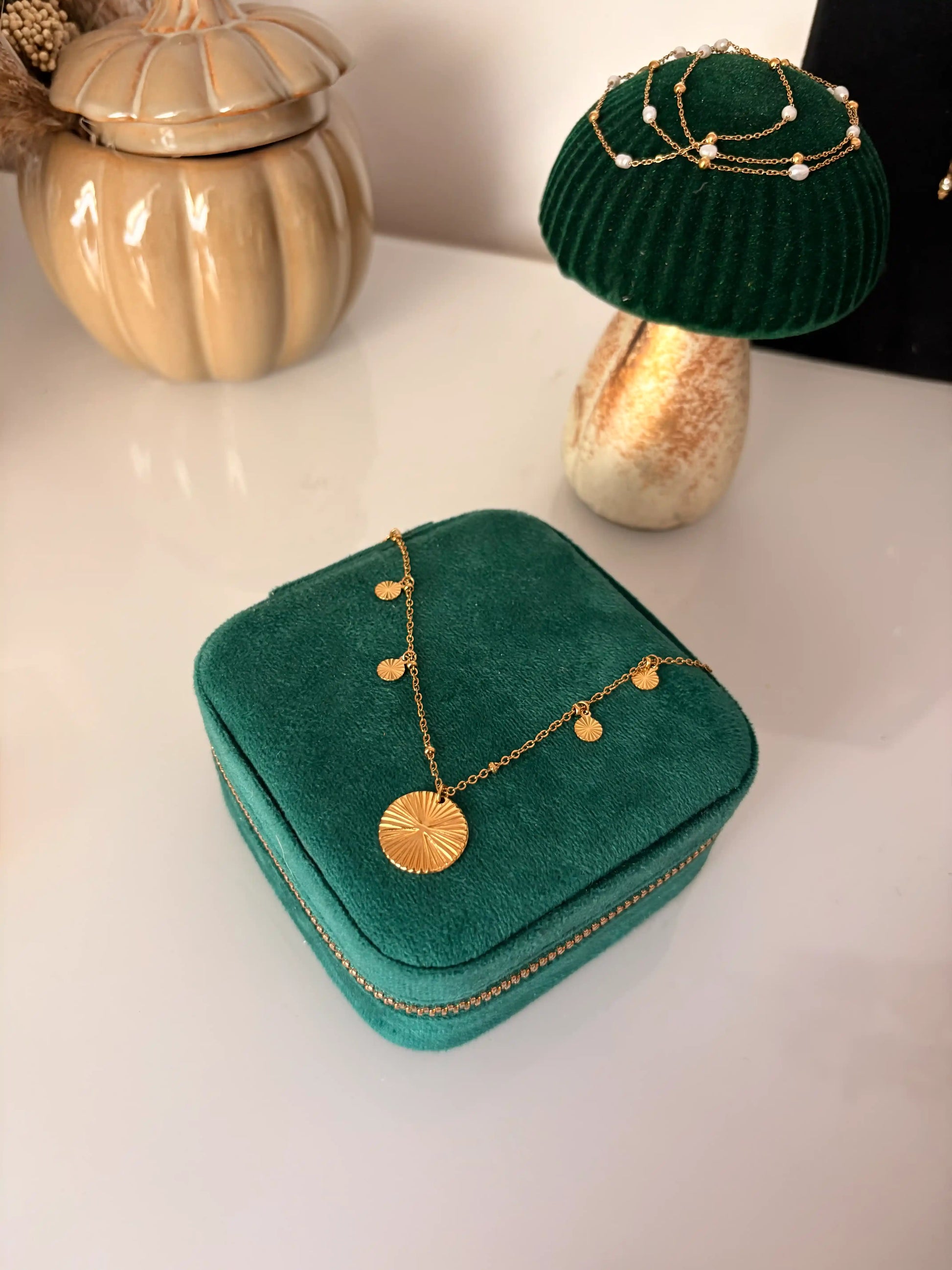 collier chaîne en acier inoxydable doré et orné d'un pendentif rond strié central et de petits sequins ronds striés sur les côtés. Bijou présenté sur une boite à bijoux vert émeraude disposée sur une table décorée d'un champignon et d'une citrouille en céramique.